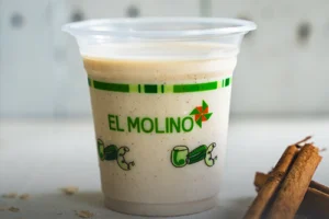 Vaso transparente con avena fría tradicional y un toque de canela, destacando el logo verde de Panadería El Molino, acompañado de astillas de canela en Cali.