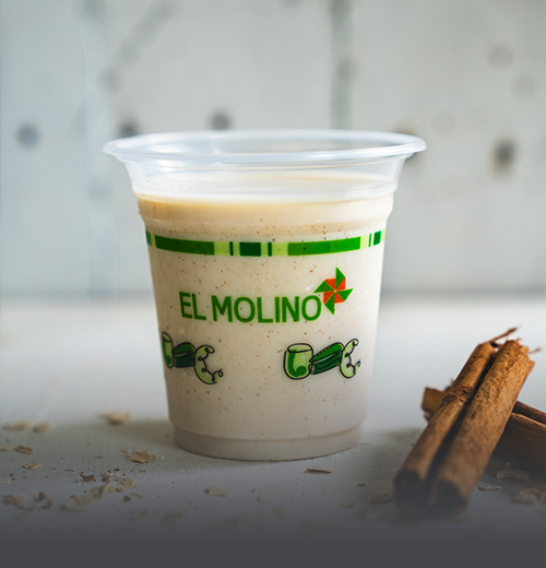 Avena Tradicional Fría - Panadería El Molino Cali Vaso transparente con avena fría tradicional y un toque de canela, destacando el logo verde de Panadería El Molino, acompañado de astillas de canela en Cali.
