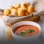 Canasta de mimbre con pan banquete artesanal dorado y redondeado, acompañado de un tazón de crema de tomate con albahaca en Panadería El Molino, Cali.