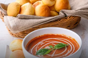 Canasta de mimbre con pan banquete artesanal dorado y redondeado, acompañado de un tazón de crema de tomate con albahaca en Panadería El Molino, Cali.