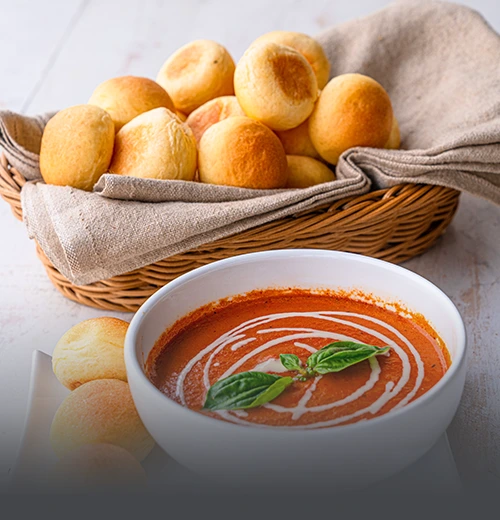 Canasta de mimbre con pan banquete artesanal dorado y redondeado, acompañado de un tazón de crema de tomate con albahaca en Panadería El Molino, Cali.