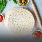 Bases de pan pizza artesanales de Panadería El Molino en Cali, rodeadas de ingredientes frescos como aceitunas, tomates cherry y salsa de tomate sobre una mesa de madera blanca con rodillo.