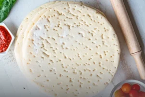 Bases de pan pizza artesanales de Panadería El Molino en Cali, rodeadas de ingredientes frescos como aceitunas, tomates cherry y salsa de tomate sobre una mesa de madera blanca con rodillo.