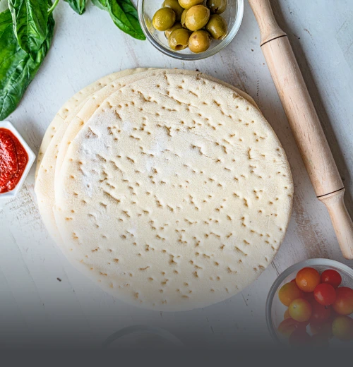 Bases de pan pizza artesanales de Panadería El Molino en Cali, rodeadas de ingredientes frescos como aceitunas, tomates cherry y salsa de tomate sobre una mesa de madera blanca con rodillo.