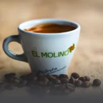 Taza de café americano caliente con logo de Panadería El Molino, rodeada de granos de café tostados sobre mesa rústica, en Cali.