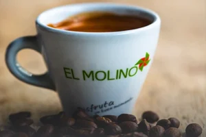 Taza de café americano caliente con logo de Panadería El Molino, rodeada de granos de café tostados sobre mesa rústica, en Cali.