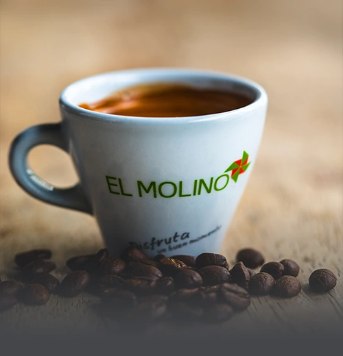 Taza de café americano caliente con logo de Panadería El Molino, rodeada de granos de café tostados sobre mesa rústica, en Cali.