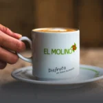 Mano sosteniendo una taza de café latte caliente con el logo de Panadería El Molino y el mensaje "Disfruta un buen momento" en Cali.