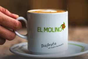 Mano sosteniendo una taza de café latte caliente con el logo de Panadería El Molino y el mensaje "Disfruta un buen momento" en Cali.