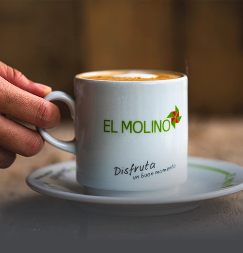 Mano sosteniendo una taza de café latte caliente con el logo de Panadería El Molino y el mensaje "Disfruta un buen momento" en Cali.