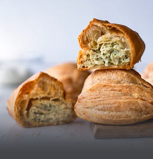 Empanada de pollo artesanal con masa dorada y crujiente, partida a la mitad mostrando su relleno de pollo desmechado sobre una tabla de madera en Panadería El Molino.