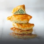 Pila de empanadas horneadas de masa hojaldrada dorada, con la de arriba partida por la mitad mostrando un abundante relleno de queso ricotta y espinaca fresca, en Panadería El Molino, Cali.