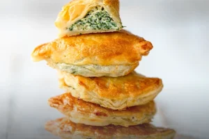 Pila de empanadas horneadas de masa hojaldrada dorada, con la de arriba partida por la mitad mostrando un abundante relleno de queso ricotta y espinaca fresca, en Panadería El Molino, Cali.