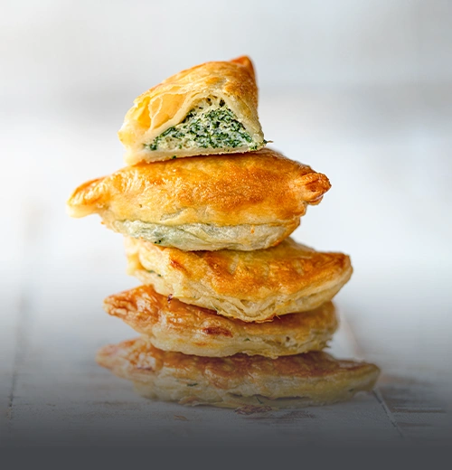 Pila de empanadas horneadas de masa hojaldrada dorada, con la de arriba partida por la mitad mostrando un abundante relleno de queso ricotta y espinaca fresca, en Panadería El Molino, Cali.
