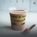 Vaso de Milo frío con la marca de Panadería El Molino, acompañado por una cuchara de madera con polvo de chocolate, en Cali.