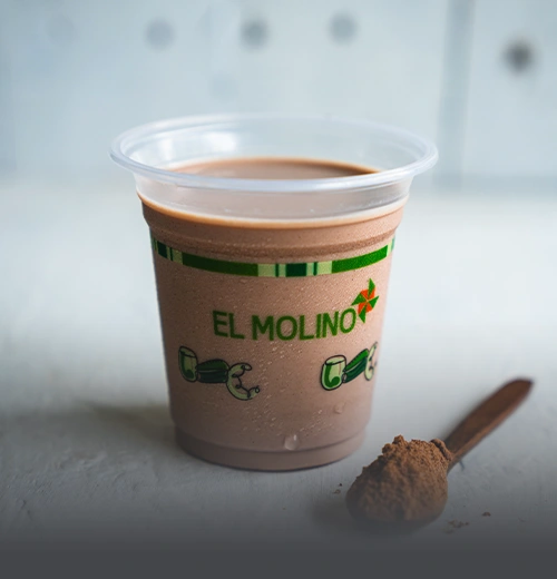Milo Frío - Panadería El Molino Cali Vaso de Milo frío con la marca de Panadería El Molino, acompañado por una cuchara de madera con polvo de chocolate, en Cali.