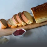 Pan aliñado de mantequilla tajado sobre una tabla de madera, acompañado de mermelada de frutos rojos y mantequilla en la mesa de Panadería El Molino.