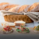 Pan baguette artesanal de corteza crujiente en canasta de mimbre y una tabla de madera con montaditos de jamón serrano y pesto en Panadería El Molino, Cali.