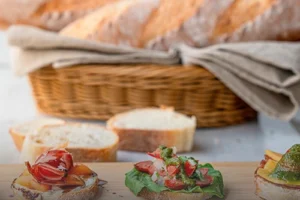 Pan baguette artesanal de corteza crujiente en canasta de mimbre y una tabla de madera con montaditos de jamón serrano y pesto en Panadería El Molino, Cali.