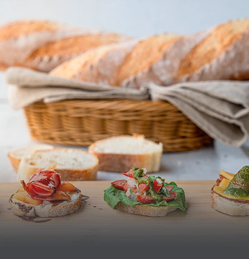 Pan baguette artesanal de corteza crujiente en canasta de mimbre y una tabla de madera con montaditos de jamón serrano y pesto en Panadería El Molino, Cali.