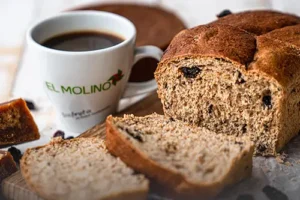Hogaza de pan hamburgo artesanal rebanado, con uvas pasas y panela, acompañado de una taza de café en Panadería El Molino, Cali.