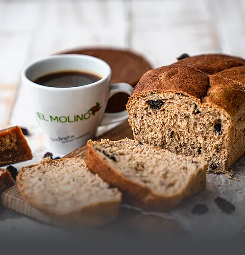 Hogaza de pan hamburgo artesanal rebanado, con uvas pasas y panela, acompañado de una taza de café en Panadería El Molino, Cali.