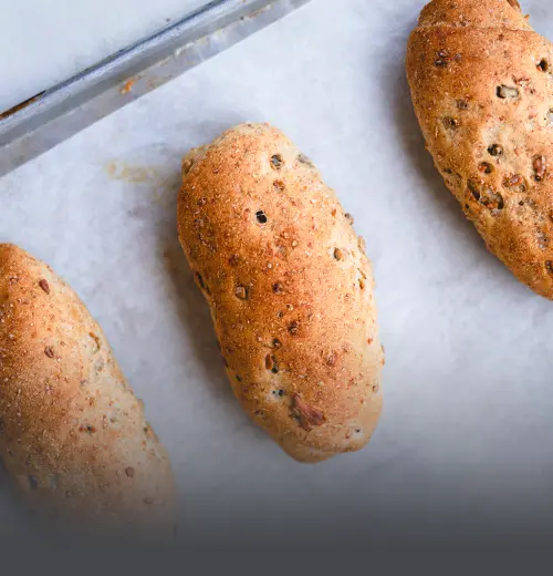 Pan Integral de Girasol y Avena - Panadería El Molino Cali Tres panes integrales artesanales cubiertos y horneados con semillas de girasol y hojuelas de avena, presentados sobre papel mantequilla en Panadería El Molino, Cali.