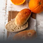Panecillos artesanales de pan integral sueco de centeno, uno de ellos cortado por la mitad para mostrar su miga suave, acompañados de naranjas frescas en Panadería El Molino, Cali.