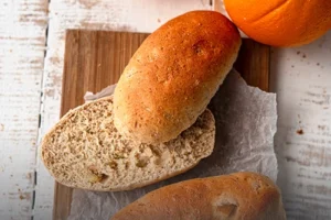Panecillos artesanales de pan integral sueco de centeno, uno de ellos cortado por la mitad para mostrar su miga suave, acompañados de naranjas frescas en Panadería El Molino, Cali.
