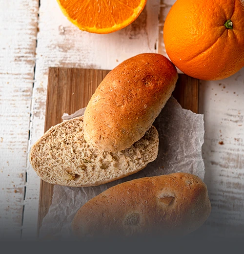 Pan Integral Sueco de Centeno - Panadería El Molino Cali Panecillos artesanales de pan integral sueco de centeno, uno de ellos cortado por la mitad para mostrar su miga suave, acompañados de naranjas frescas en Panadería El Molino, Cali.