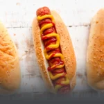 Pan artesanal para perros calientes cubierto con ajonjolí, mostrado en una preparación clásica con salchicha y salsas en Panadería El Molino, Cali.