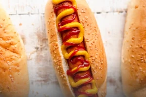 Pan artesanal para perros calientes cubierto con ajonjolí, mostrado en una preparación clásica con salchicha y salsas en Panadería El Molino, Cali.