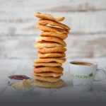 Alta pila de tostadas dulces artesanales y crujientes, acompañadas de porciones de mantequilla, mermelada y una taza de café caliente con el logo de Panadería El Molino, Cali.
