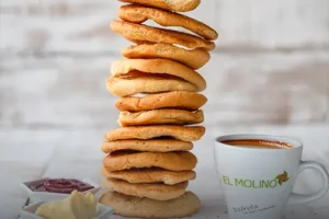 Alta pila de tostadas dulces artesanales y crujientes, acompañadas de porciones de mantequilla, mermelada y una taza de café caliente con el logo de Panadería El Molino, Cali.