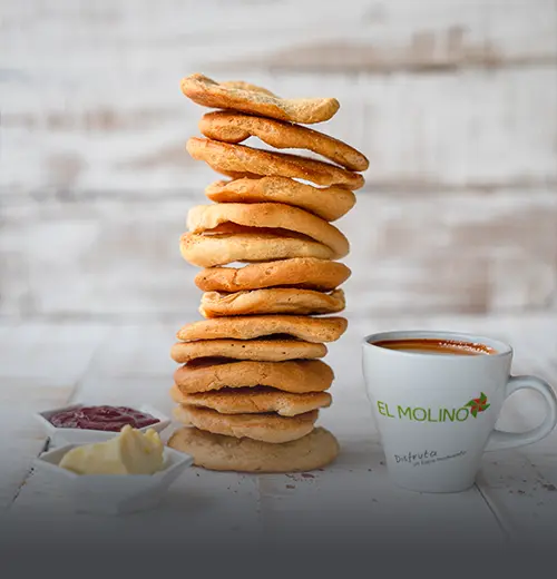 Alta pila de tostadas dulces artesanales y crujientes, acompañadas de porciones de mantequilla, mermelada y una taza de café caliente con el logo de Panadería El Molino, Cali.