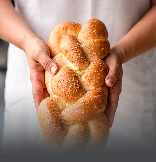 Trenza Aliñada Artesanal - Panadería El Molino Cali Trenza aliñada artesanal recién horneada, sostenida en las manos de un panadero y cubierta con ajonjolí en Panadería El Molino, Cali.