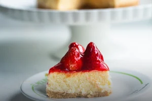 Porción de cheesecake artesanal con cobertura de fresas brillantes, servida en un plato con el logo de Panadería El Molino, con el cheesecake entero al fondo desenfocado.