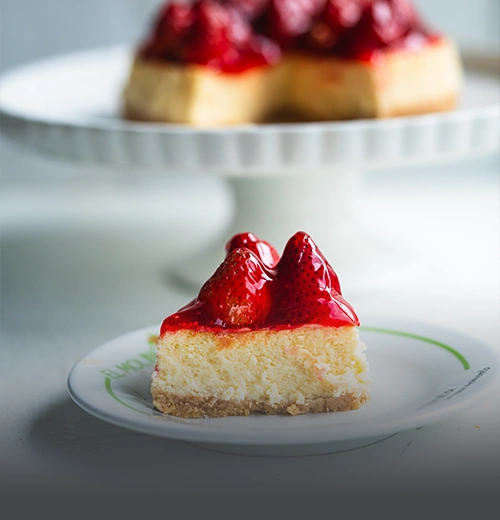 Porción de Cheesecake de Fresas - Panadería El Molino Cali Porción de cheesecake artesanal con cobertura de fresas brillantes, servida en un plato con el logo de Panadería El Molino, con el cheesecake entero al fondo desenfocado.