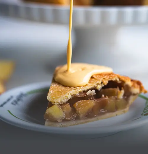 Primer plano de una porción de pie de manzana artesanal con salsa cremosa de vainilla en un plato blanco de Panadería El Molino, con manzanas frescas al fondo.