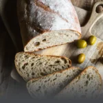 Hogaza de pan de masa madre con olivas verdes, cortada en rebanadas sobre una tabla de madera rústica, resaltando la miga con trozos de aceituna en Panadería El Molino.