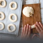 Manos moldeando masa fresca de pandebono tradicional sobre una tabla rústica de madera, junto a una bandeja lista para entrar al horno, de Panadería El Molino, Cali.
