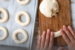 Manos moldeando masa fresca de pandebono tradicional sobre una tabla rústica de madera, junto a una bandeja lista para entrar al horno, de Panadería El Molino, Cali.