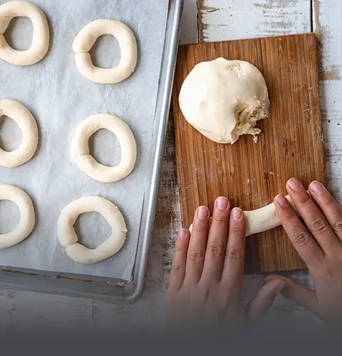 Masa de Pandebono para Hornear en Casa - Panadería El Molino Cali Manos moldeando masa fresca de pandebono tradicional sobre una tabla rústica de madera, junto a una bandeja lista para entrar al horno, de Panadería El Molino, Cali.