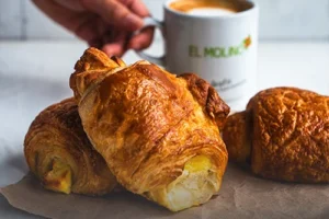 Croissant de queso hojaldrado artesanal con queso dorado en la corteza, sobre una mesa de madera rústica en Panadería El Molino, Cali.