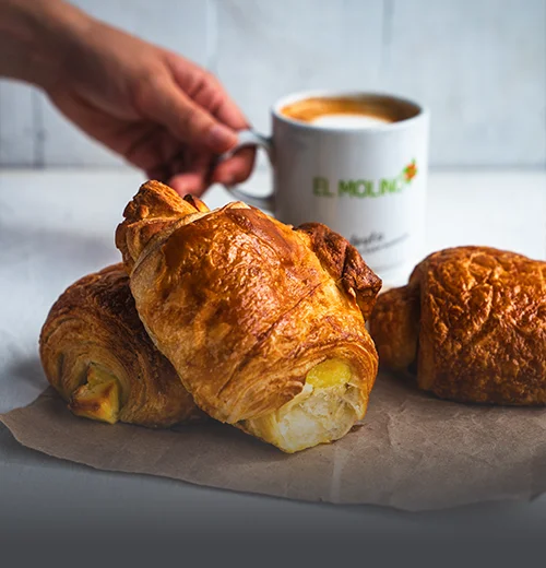 Croissant de queso hojaldrado artesanal con queso dorado en la corteza, sobre una mesa de madera rústica en Panadería El Molino, Cali.