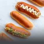 Panes artesanales para perro caliente recién horneados sobre una mesa blanca, algunos decorados con salsas y aderezos en Cali.