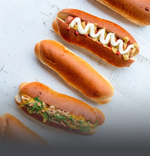 Panes artesanales para perro caliente recién horneados sobre una mesa blanca, algunos decorados con salsas y aderezos en Cali.