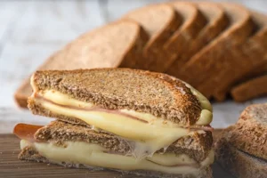 Sándwich tostado de queso derretido y jamón preparado con pan tajado integral de grano entero, junto a varias rebanadas al fondo sobre tabla de madera en Cali.