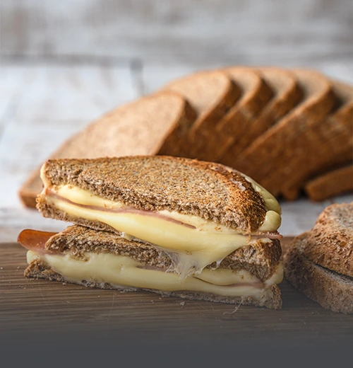 Sándwich tostado de queso derretido y jamón preparado con pan tajado integral de grano entero, junto a varias rebanadas al fondo sobre tabla de madera en Cali.