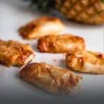Pasteles hawaianos de hojaldre crujiente recién horneados, rellenos de jamón, queso y piña, acompañados de una piña natural al fondo en Cali.